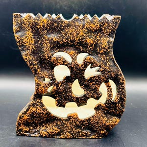 May include: A Halloween-themed ceramic candle holder shaped like a jack-o'-lantern. The holder is black with orange speckles and features cut-out eyes, nose, and a smiling mouth. The interior is white, designed to hold a candle.