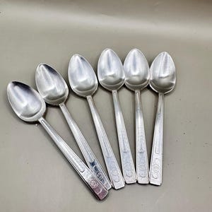 May include: A set of six silver-toned metal spoons arranged in a fan pattern. Each spoon features a smooth, oval-shaped bowl and a straight handle decorated with vertical grooved lines and a circular embossed floral motif near the base of the handle.