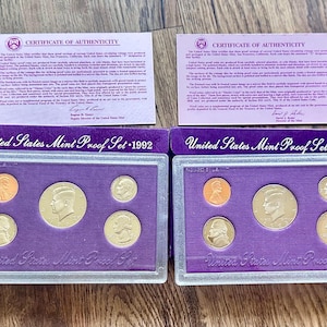 Op de afbeelding: Twee United States Mint Proof Sets, 1992 en 1993, elk met vijf munten in een paarse koffer. De sets bevatten een penny, een halve dollar en andere munten. Elke set heeft een certificaat van echtheid.