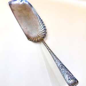 May include: Silver-toned cake server with a wide, rectangular blade and a decorative, ornate handle. The blade has a scalloped edge and a detailed, swirling design along the side. The handle features intricate floral and figure engravings.