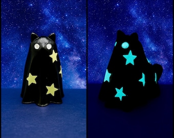 以下が含まれることがあります： 黄色い星の切り抜きが付いた黒い猫の形をした置物。照明されると、置物はターコイズ色の光で光ります。背景は星空の濃い青色です。