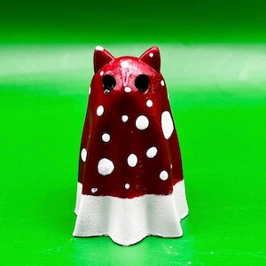 Può includere: Una stravagante statuetta a forma di fantasma in ceramica fatta a mano con un volto simile a quello di un gatto. Il fantasma è dipinto di rosso intenso con pois bianchi e un bordo inferiore bianco. La statuetta ha fori neri per gli occhi e piccole orecchie a punta.
