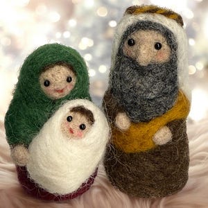 Op de afbeelding: Drie naaldgevilte figuren die een kersttafereel voorstellen. Eén figuur draagt een groene kap en houdt een kleine baby vast die in witte wol is gewikkeld. Daarnaast staat een groter figuur met een grijze baard, gekleed in een bruine mantel.