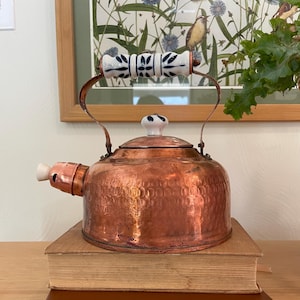 Puede incluir: Una tetera de cobre con acabado martillado, apoyada sobre dos libros. La tetera tiene un asa con diseño blanco y azul y un pico blanco. Los libros son marrones y beige. El fondo incluye una impresión botánica enmarcada.