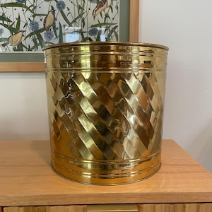 May include: A cylindrical, gold-colored metal wastebasket with a diagonal, textured pattern. The wastebasket is sitting on a light-colored wooden cabinet. A framed botanical print hangs in the background.