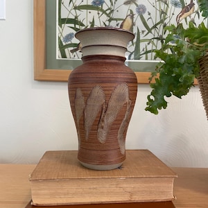 Puede incluir: Un jarrón de cerámica marrón con un diseño texturizado, colocado sobre dos libros. El jarrón tiene un borde beige claro y un cuerpo marrón oscuro con un patrón tallado. Los libros están apilados, con una mesa de madera en primer plano. El jarrón mide unos 25 cm de alto.