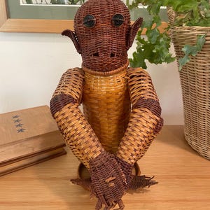 May include: A handcrafted wicker sculpture of a humanoid figure with a brown head, ears, and arms, and a tan torso. The sculpture has button eyes and is posed with its arms crossed. The sculpture is on a wooden surface.