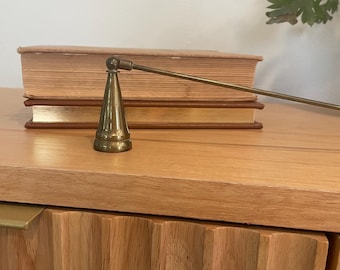 May include: A brass candle snuffer with a conical base and long handle rests on a wooden surface. Two books with brown covers are behind the snuffer. The wooden cabinet has a fluted design.