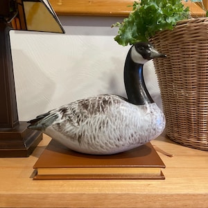 Puede incluir: Una figura de ganso de cerámica con cabeza y cuello negros, y cuerpo blanco y gris. El ganso está posado sobre un libro marrón. La figura es un objeto decorativo.