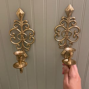 May include: Two matching wall-mounted candle holders crafted from polished brass. Each piece features an elongated, symmetrical scrollwork design with a diamond-shaped top and a circular base plate supporting a central cup for holding a candle.