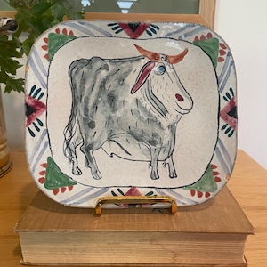 Puede incluir: Plato de cerámica pintado a mano con una ilustración de un toro. El toro es gris con cuernos rojos y un ojo azul, sobre un fondo crema. El plato tiene forma cuadrada con un borde decorativo con detalles azules, rojos y verdes. El plato se exhibe sobre un soporte dorado.
