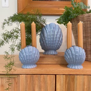Può includere: Composizione decorativa su un mobile in legno. Presenta tre portacandele a forma di conchiglia azzurra, tre candele beige e una scultura di conchiglia più grande. Sono presenti anche un cesto di vimini e del verde.