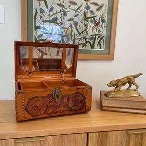 May include: An open, vintage leather jewelry box with a floral embossed design. The box has a mirrored lid and is sitting on a wooden surface. A gold-colored dog statue rests on a stack of books.