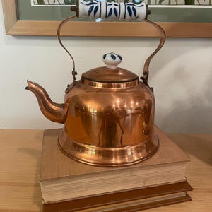 May include: A polished copper tea kettle with a curved spout and a rounded body. The handle features a white ceramic grip with blue painted leaf patterns, and the lid is topped with a matching white and blue ceramic knob finial.