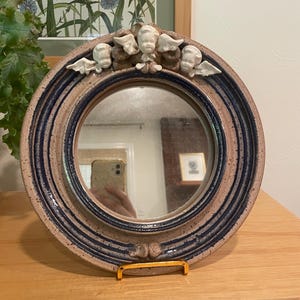 May include: A round ceramic mirror with a blue and tan rim. The rim features a raised design with four small angel figures. The mirror is supported by a gold-colored metal stand. The mirror reflects the room.