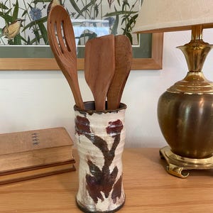 May include: A cylindrical ceramic utensil holder with a cream-colored glaze and dark brown abstract brushstroke patterns. Three wooden kitchen utensils, including a slotted spoon and two flat spatulas, are placed inside the vessel.