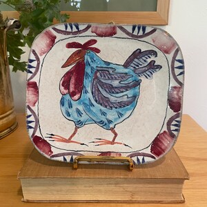 May include: Square ceramic plate with a hand-painted rooster design. The rooster is blue with red accents, set against a cream background. The border features a repeating pattern of red and blue shapes. The plate is displayed on a book.