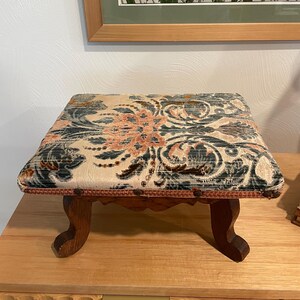 Könnte beinhalten: Ein kleiner rechteckiger Fußhocker mit gepolsterter Sitzfläche, die mit einem strukturierten Stoff mit Blumenmuster in Pfirsich-, Blaugrün- und Cremetönen bezogen ist. Das Untergestell besteht aus dunkelbraunem Holz mit vier geschwungenen Beinen.