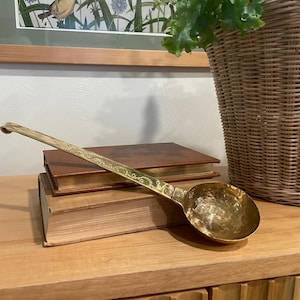 May include: A vintage brass ladle with a hammered bowl and ornate handle rests on two antique books. The ladle is gold-colored and appears to be made of brass. The books are brown and have aged pages.