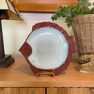 May include: A ceramic fish-shaped plate with a white center and a reddish-brown border. The plate is displayed on a gold-colored stand, showcasing its unique design. The plate is a decorative item.