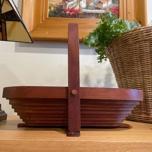 Puede incluir: Una cesta de madera marrón rojizo con asa. La cesta tiene un diseño escalonado y está colocada sobre una superficie de madera. Una lámpara decorativa y una cesta de mimbre están en el fondo. El diseño de la cesta es único.