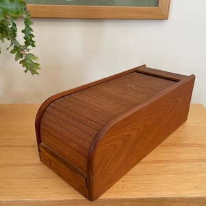 Puede incluir: Una panera de madera con tapa enrollable. La caja es de madera de tonos cálidos y tiene forma rectangular con bordes redondeados. La tapa enrollable está hecha de finas láminas de madera.