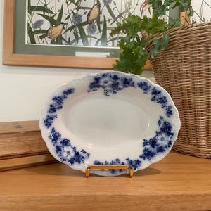 May include: An oval, white ceramic serving dish with a scalloped edge and blue floral pattern. The dish is displayed on a gold-colored metal stand. A stack of books and a woven basket are in the background.