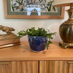 Puede incluir: Una maceta de cerámica azul con un diseño en relieve texturizado que contiene una planta de hojas verdes. La maceta está situada entre una figura de perro de latón sobre dos libros apilados y una lámpara de mesa de latón con pantalla beige.