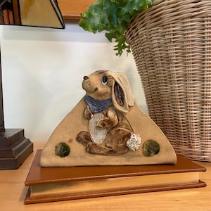 May include: A ceramic bunny figurine with a blue scarf and a carrot. The bunny is light brown with dark brown accents and is sitting on a tan triangular base. The base has two green accents and is resting on a brown book.