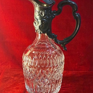 Puede incluir: Una jarra de cristal transparente con asa y tapa de metal plateado. La jarra tiene un patrón tallado en diamante y se encuentra sobre una superficie roja. El asa y la tapa presentan detalles ornamentados.