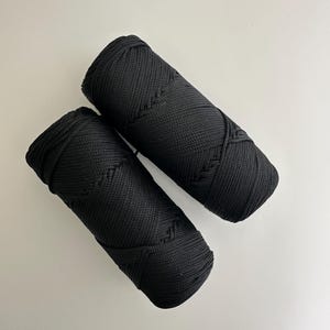 May include: Two black macrame cord spools are shown against a white backdrop. The tightly wound cord displays a woven texture. The cylindrical spools have a decorative pattern on the ends. Ideal for craft projects, the image highlights the cord's texture and form.