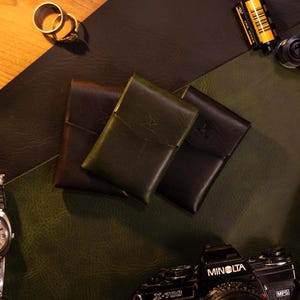 May include: Three leather cardholders in shades of brown and green are arranged on a textured surface. The cardholders have a simple design with a flap closure. A Minolta X-700 camera and film rolls are also visible.