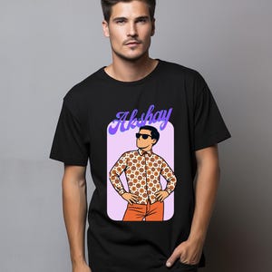 May include: Black t-shirt featuring a graphic of a man in a patterned shirt, orange pants, and sunglasses. The graphic is inside a purple rectangle with the name "Akshay" in cursive.