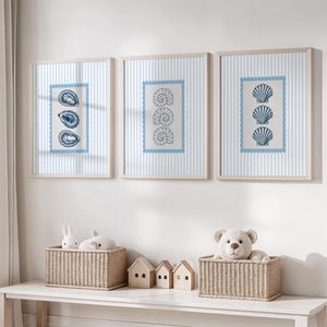 May include: Three framed prints with a nautical theme. Each print features a different seashell design in blue against a striped background. The frames are light wood. Below, two wicker baskets hold stuffed animals and miniature wooden houses.