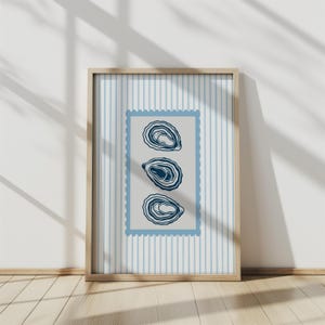 May include: A framed print featuring three stylized oyster illustrations in navy blue. The artwork is set against a light gray background within a blue postage stamp-like border, and a light blue and white striped background. The frame is made of light wood.