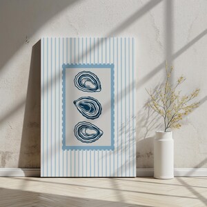 May include: A canvas print featuring three stylized oyster illustrations in a blue frame. The background has vertical blue stripes. The artwork is displayed on a wooden floor, leaning against a white wall.