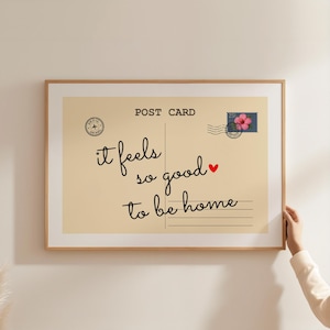 Puede incluir: Impresión enmarcada de postal con las palabras "it feels so good to be home" en escritura cursiva. La postal tiene una estética vintage con un sello y un pequeño corazón rojo.