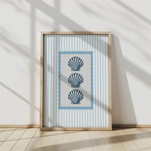 May include: A framed print featuring three stylized blue seashell illustrations on a light gray background within a blue postage stamp border. The background has vertical light blue stripes. The frame is made of light-colored wood.