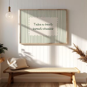 May include: A framed print with a light wood frame hangs on a white wall. The print features a green and white striped background with a cream-colored center that reads "Take a seat sweet cheeks" in green script. A bench with a cream-colored cushion sits below.
