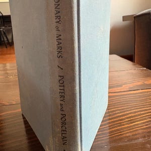 May include: A light blue book titled "Dictionary of Marks / Pottery and Porcelain" with the word "Crown" at the bottom. The book is standing upright on a wooden surface.