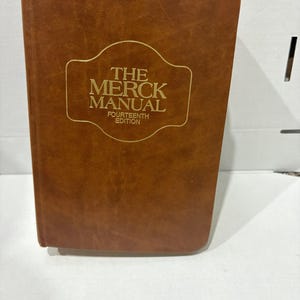May include: A brown leather-bound book titled "The Merck Manual, Fourteenth Edition" with gold lettering. The book has a rectangular shape and appears to be in good condition. The cover has a slightly textured appearance.