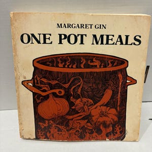 Puede incluir: Libro de cocina vintage titulado "ONE POT MEALS" de Margaret Gin. La portada presenta una olla marrón con ilustraciones naranjas y rojas de verduras. El libro tiene un aspecto desgastado y envejecido.