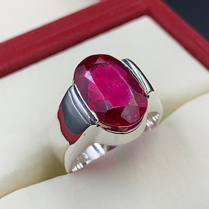 May include: A silver ring featuring a large, oval-cut ruby gemstone. The ruby is a deep, vibrant red color and is set in a polished silver band. The ring is displayed in a red jewelry box.
