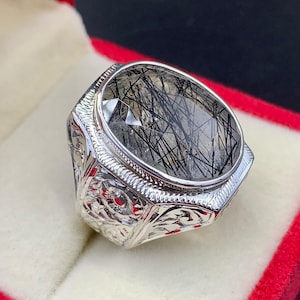 Pode incluir: Anel de prata com uma pedra preciosa de corte oval. A pedra é transparente com inclusões pretas em forma de agulha. O anel tem um design ornamentado e gravado na banda.