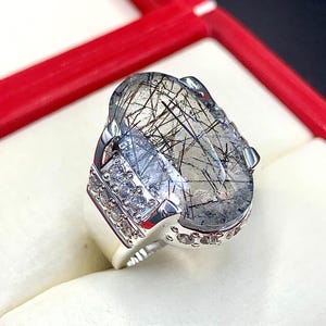 Pode incluir: Anel de prata com uma grande pedra preciosa oval com inclusões pretas. O anel tem um design moderno com uma banda adornada com pequenas pedras transparentes. A pedra preciosa é engastada em prata polida.