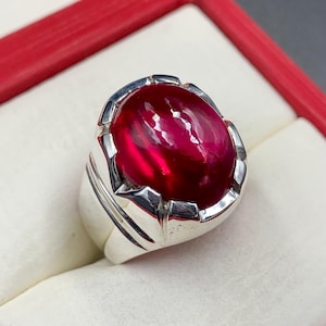 Op de afbeelding: Een zilveren ring met een grote, ovale, dieprode edelsteen. De edelsteen is gezet in een gepolijste zilveren band met decoratieve details. De ring wordt getoond tegen een witte achtergrond, met een rode rand zichtbaar.
