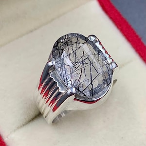Pode incluir: Um anel de prata com uma grande pedra preciosa oval com inclusões pretas em forma de agulha. O anel tem uma banda metálica polida com um design canelado nas laterais. A pedra preciosa é transparente com padrões lineares escuros.