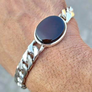 May include: A silver chain bracelet with a large oval-shaped black gemstone. The bracelet has a curb chain design.