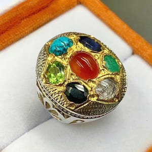 Puede incluir: Un anillo de plata con engaste dorado, adornado con piedras preciosas de colores. El diseño del anillo incluye una piedra roja central rodeada de gemas turquesas, verdes, azules y negras. El anillo se presenta en una caja de terciopelo naranja.