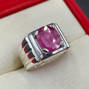 May include: A silver ring with a large, oval-cut ruby gemstone. The ruby is a deep pinkish-red colour and is set in a rectangular silver band with a ridged design on the sides. The ring is displayed in a red box.